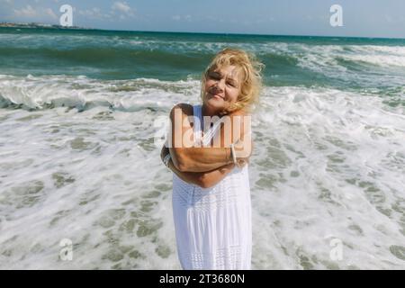 Donna anziana sorridente che si abbraccia vicino al mare in spiaggia Foto Stock