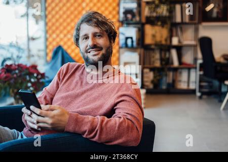 Uomo sorridente con smartphone seduto a casa Foto Stock