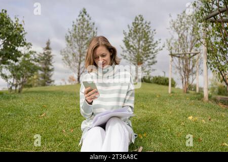 Donna seduta sull'erba e che usa lo smartphone nel parco Foto Stock