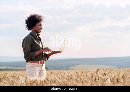 Sorridente agricoltore afro con un computer portatile tra le colture di orzo nei campi Foto Stock