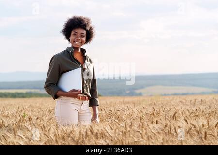 Un lavoratore afro sorridente con un computer portatile tra le colture di orzo nei campi Foto Stock