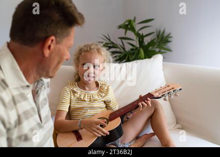Ragazza sorridente che suona la chitarra seduta accanto al padre nel salotto di casa Foto Stock