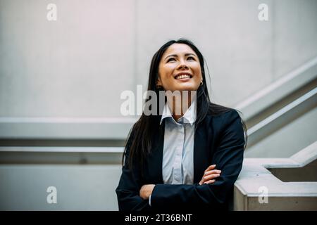 Donna d'affari sorridente con le braccia incrociate che si appoggiano alla ringhiera delle scale che guarda in alto all'ufficio Foto Stock