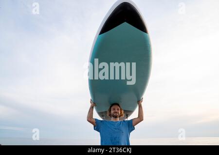 Uomo sorridente che porta il paddleboard sotto il cielo al tramonto Foto Stock