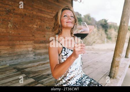 Donna sorridente che si gode un bicchiere di vino seduto su una veranda di legno Foto Stock