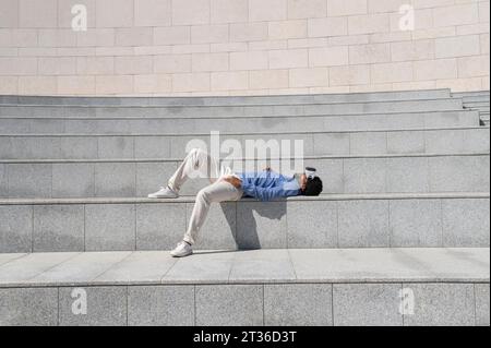 Uomo spensierato che indossa un simulatore di realtà virtuale e si sdraia sui gradini Foto Stock