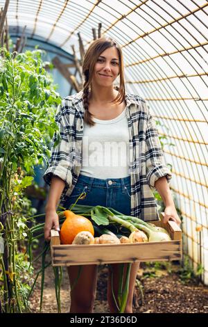 Giovane lavoratore agricolo sorridente con verdure appena raccolte in cassa in serra Foto Stock