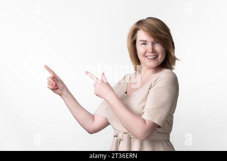 Donna bionda europea matura adulta vestita con cintura allacciata davanti alla fotocamera. La femmina punta le dita verso l'alto su qualcosa di interessante Foto Stock