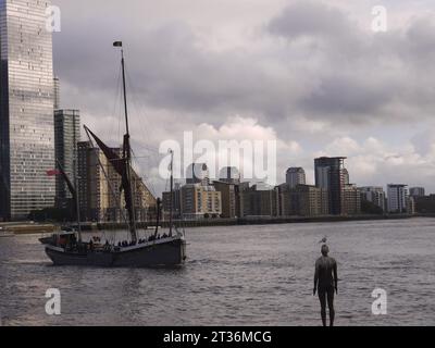 Limehouse, Londra, Regno Unito - 21 ottobre 2023: Antony Gormley's Another Time, con un gabbiano arroccato sulla testa, guarda passare la chiatta a vela del Tamigi. Foto Stock