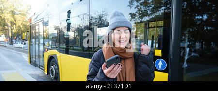 Persone moderne e stile di vita. Felice ragazza asiatica urla da gioia, festeggia, si trova vicino ai trasporti pubblici degli autobus e sembra stupito Foto Stock