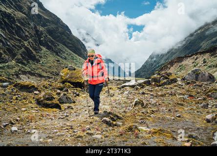 Donna con occhiali da sole con zaino e bastoncini da trekking vestita di giacca softshell rossa durante il trekking nel Parco Nazionale Makalu Barun in Nepal. Escursione in montagna Foto Stock