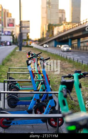 Scooter elettrici colorati e a pagamento in un parcheggio sul marciapiede. Sullo sfondo, una vista leggermente sfocata delle strade della città. Foto Stock