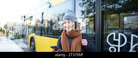 Ritratto di ragazza in piedi vicino a un autobus in una fermata, in attesa del suo trasporto pubblico, controlla il calendario su smartphone applicazione, tiene il telefono cellulare, indossa Foto Stock