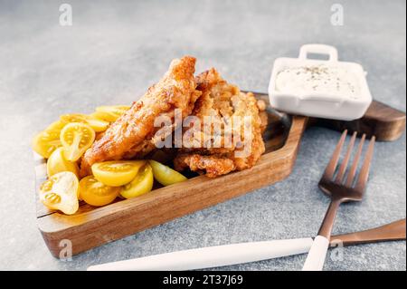 Il piatto di pesce è un filetto di merluzzo fritto con una salsa cremosa e pomodori di prugne gialli, servito su un piatto di legno a forma di pesce. Foto Stock