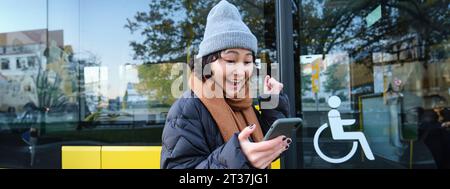 Persone moderne e stile di vita. Felice ragazza asiatica urla da gioia, festeggia, si trova vicino ai trasporti pubblici degli autobus e sembra stupito Foto Stock