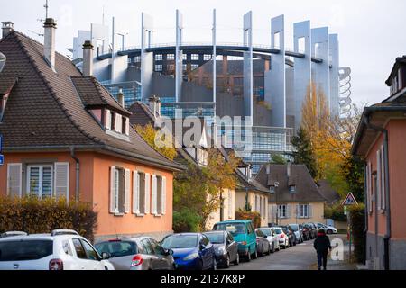 Strasburgo, Francia - 22 novembre 2023: Le tradizionali case arancioni con tetti in tegole sono in primo piano, con il torreggiante Parlamento europeo di Strasburgo visibile sullo sfondo - pedoni e automobili Foto Stock