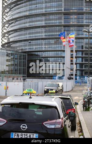 Strasburgo, Francia - 22 novembre 2023: Taxi in attesa in coda con un edificio a spirale a più livelli del Parlamento sullo sfondo, accompagnati da bandiere nazionali. Foto Stock