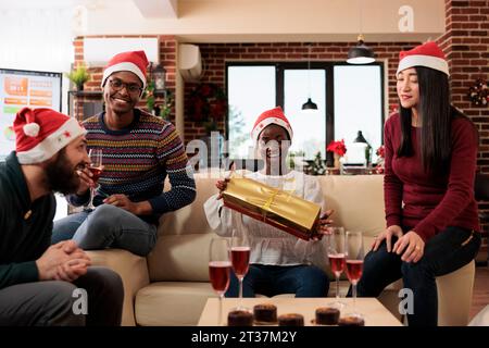 Felice donna afro-americana che tiene una confezione regalo natalizia dai colleghi mentre si siede sul divano in ufficio. Diversi colleghi che condividono regali e bevono alla festa aziendale di natale Foto Stock