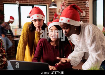 Giovani donne colleghi che indossano cappelli di babbo natale discutono e collaborano alle attività a natale. Diversi colleghi sorridenti lavorano insieme su un computer portatile in un ufficio decorato la vigilia di Natale Foto Stock