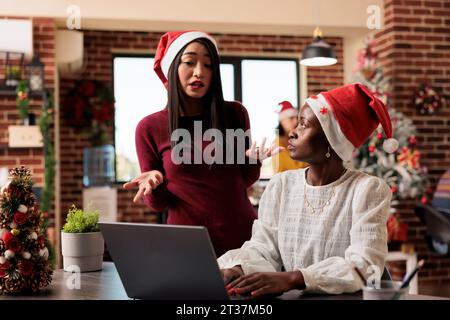 Le donne che collaborano a babbo natale discutono e discutono su un laptop in un'area di lavoro decorata. Colleghi asiatici e afroamericani che fanno lavoro di squadra nello spazio di coworking invernale Foto Stock