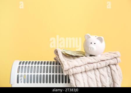 Salvadanaio con coperta calda e denaro sul riscaldatore elettrico convettoriale vicino alla parete gialla a casa. Concetto di risparmio termico Foto Stock