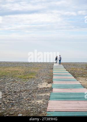 Una coppia che si tiene per mano cammina lungo il sentiero verso il mare. Amanti sullo sfondo del mare. Sagome di persone. Strada fatta di pallet di legno Foto Stock