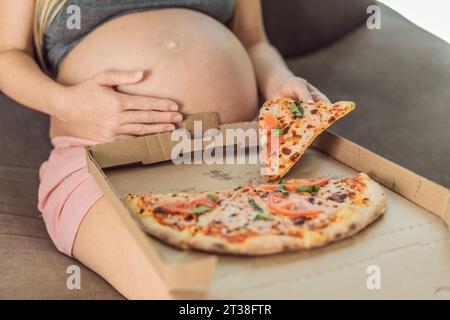 Una donna incinta si diverte a gustare una fetta di pizza, assaporando un momento di indulgenza e soddisfacendo il suo desiderio di una delizia deliziosa e confortante. Entusiasta Foto Stock