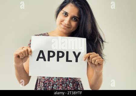 La donna con gli occhi espressivi e i capelli scuri che scorrono tiene con sicurezza un cartello con la scritta "APPLY" (APPLICA). Il motivo floreale del suo abbigliamento contrasta con la semplice clea Foto Stock