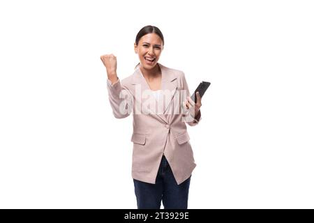 giovane donna d'affari gioiosa con un taglio di capelli a coda di cavallo vestita con una giacca beige regge un telefono Foto Stock