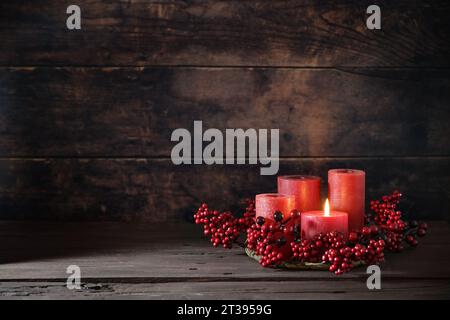 Frist Advent con decorazione a bacche rosse e candele in una corona, una e' illuminata, decor per la casa delle vacanze su uno sfondo rustico scuro di legno, spazio copia Foto Stock