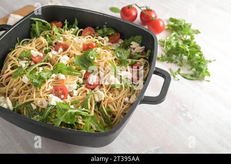 Insalata di spaghetti con pomodori, rucola, mozzarella e olive in un'ampia ciotola nera, pasto mediterraneo freddo su un tavolo dipinto di bianco Foto Stock
