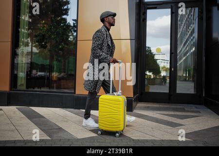 Un modello maschile elegantemente vestito cammina lungo le strade cittadine con una valigia gialla in mano. Uomo nero turista all'aperto vicino all'edificio dell'hotel. Uomo afro-americano Foto Stock