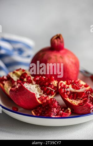 Primo piano dei semi di melograno rossi maturi su un piatto rustico bianco con bordo blu su sfondo grigio. Foto Stock