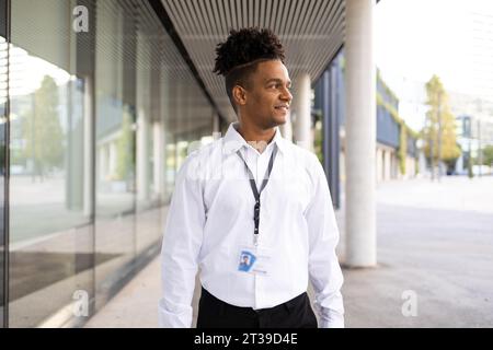 Sicuro lavoratore afroamericano con abiti formali e dreadlock ritorti rasati in piedi accanto a un edificio moderno sulla strada che guarda lontano Foto Stock