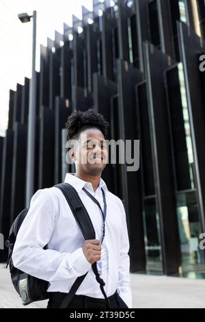 Vista laterale del ritratto di un giovane uomo afro-americano in camicia bianca che guarda la macchina fotografica mentre cammini vicino all'edificio moderno sulla strada della città Foto Stock