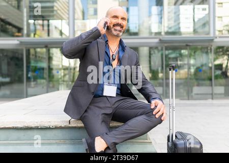 Ritratto di un uomo d'affari che parla al telefono per strada vicino alla sua valigia Foto Stock