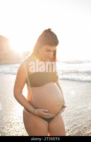 Felice donna incinta in bikini che guarda in basso e tocca la pancia mentre si trova in piedi contro il mare al tramonto Foto Stock