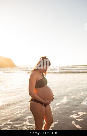 Vista laterale di una donna felice incinta in bikini che guarda in basso e tocca la pancia mentre si stende contro il mare al tramonto Foto Stock