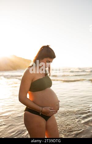 Vista laterale di una donna felice incinta in bikini che guarda in basso e tocca la pancia mentre si stende contro il mare al tramonto Foto Stock