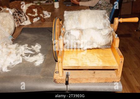 Un carder in legno a mano posto su un tavolo con soffice lana bianca, circondato da materiali per maglieria e artigianali. Foto Stock