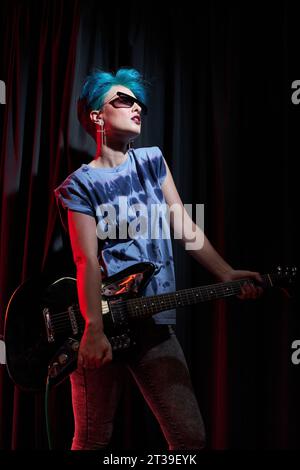 Giovane musicista professionista in capelli tinti e abiti casual con occhiali da sole che portano la chitarra in mano mentre si esibiscono sul palco Foto Stock