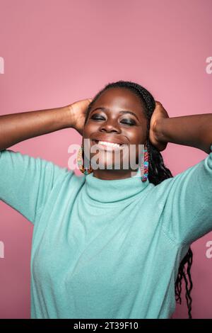 Una gioiosa donna africana con i capelli intrecciati, le mani sulla testa, sorridente, decorata con orecchini colorati e un maglione turchese a dolcevita Foto Stock