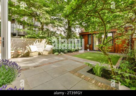 Area sul retro con sedie su patio piastrellato e piccolo giardino con capannone di legno circondato da alberi e piante lussureggianti Foto Stock