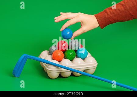 Ravvicinare la mano del raccolto tenendo la palla blu sopra la scatola da golf con palline colorate e posizionate su una superficie verde in uno studio moderno Foto Stock