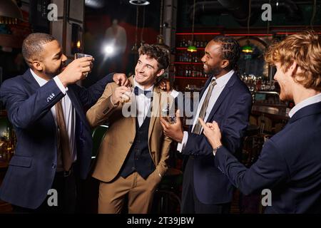 bravi sposi interrazziali indossati in modo formale si congratulano con un amico al bar, uomini con bicchieri di whisky Foto Stock