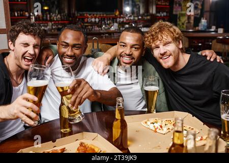 gruppo di amici multietnici felici che abbracciano e brindano con un bicchiere di birra, trascorrendo del tempo libero al bar Foto Stock