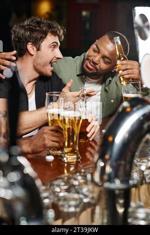 afroamericano che abbraccia un amico eccitato chiacchierando e tenendo birra al bar, amicizia maschile Foto Stock