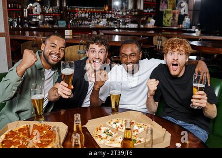 gruppo di amici interrazziali entusiasti che fanno il tifo con bicchieri di birra al bar, uomini alla festa di scapolo Foto Stock