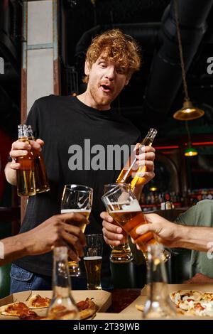 uomo con capelli ricci che regge biberon con birra vicino agli amici che tosta bicchieri al bar Foto Stock
