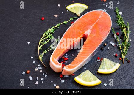 Bistecca di pesce fresco crudo con ingredienti da cucina, erbe aromatiche e limone su sfondo nero, vista dall'alto. Bistecca di salmone con timo e limone su tavola nera. Foto Stock